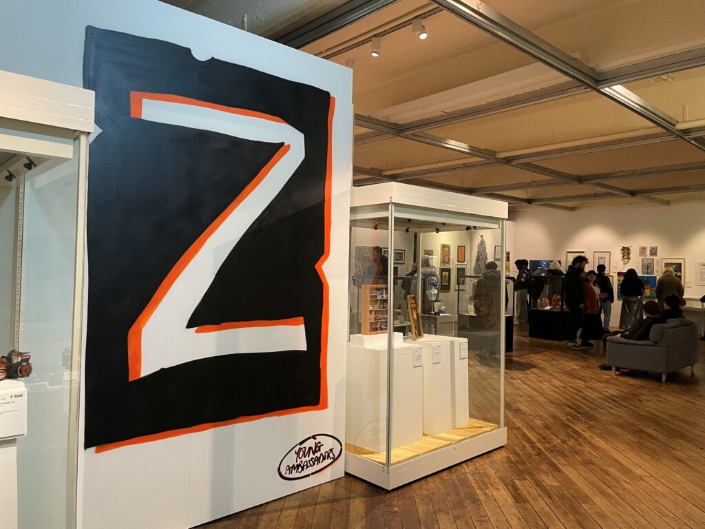An art gallery interior with wooden floors and white walls. In the foreground, a large black and orange mural featuring a bold letter “Z” with the words “Young Ambassadors” at the bottom is displayed on a partition wall. Next to it is a glass display case containing several small artworks or objects on white plinths. In the background, more framed artworks are hung on the walls, and several people are viewing the exhibits. The space is well-lit with ceiling track lights.