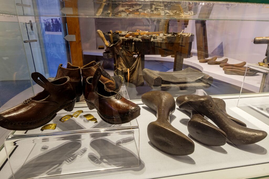 A museum display case containing several pairs of traditional leather shoes alongside wooden shoe lasts. Behind the case is a workbench with historic shoemaking tools, creating a scene that highlights traditional cobbling craftsmanship.