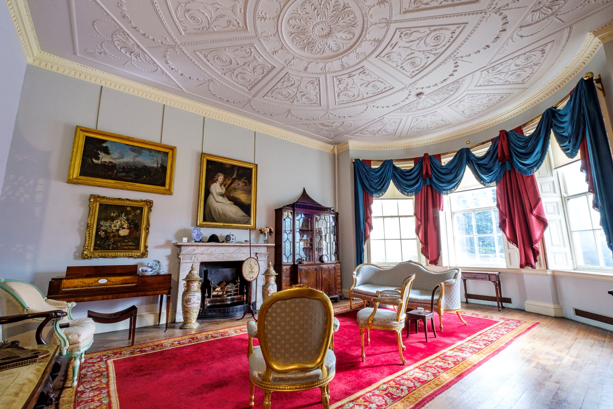 a view of a georgian room, high ceiling, large bay window and delicate wooden furniture Walls are blue, carved ceiling is white