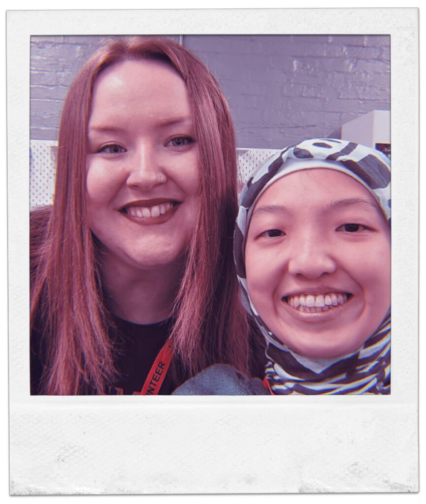 A Polaroid‑style photograph showing two people sitting close together indoors. One has long straight hair and is wearing a lanyard. The background includes a brick wall and patterned fabric panels.