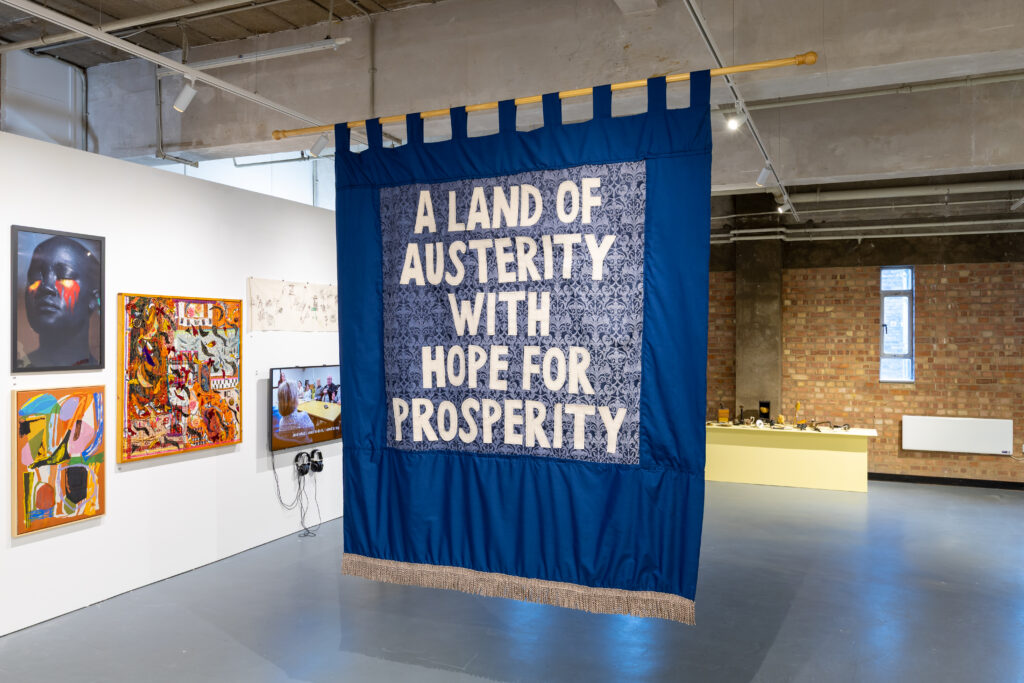 A large blue and patterned textile banner hanging in a gallery space, with bold text reading “A LAND OF AUSTERITY WITH HOPE FOR PROSPERITY” displayed in the centre.