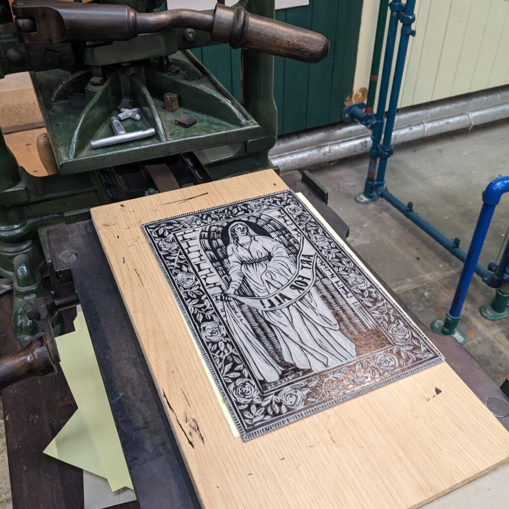 A carved printing block resting on a vintage press bed, depicting a robed figure framed by intricate floral and architectural patterns.