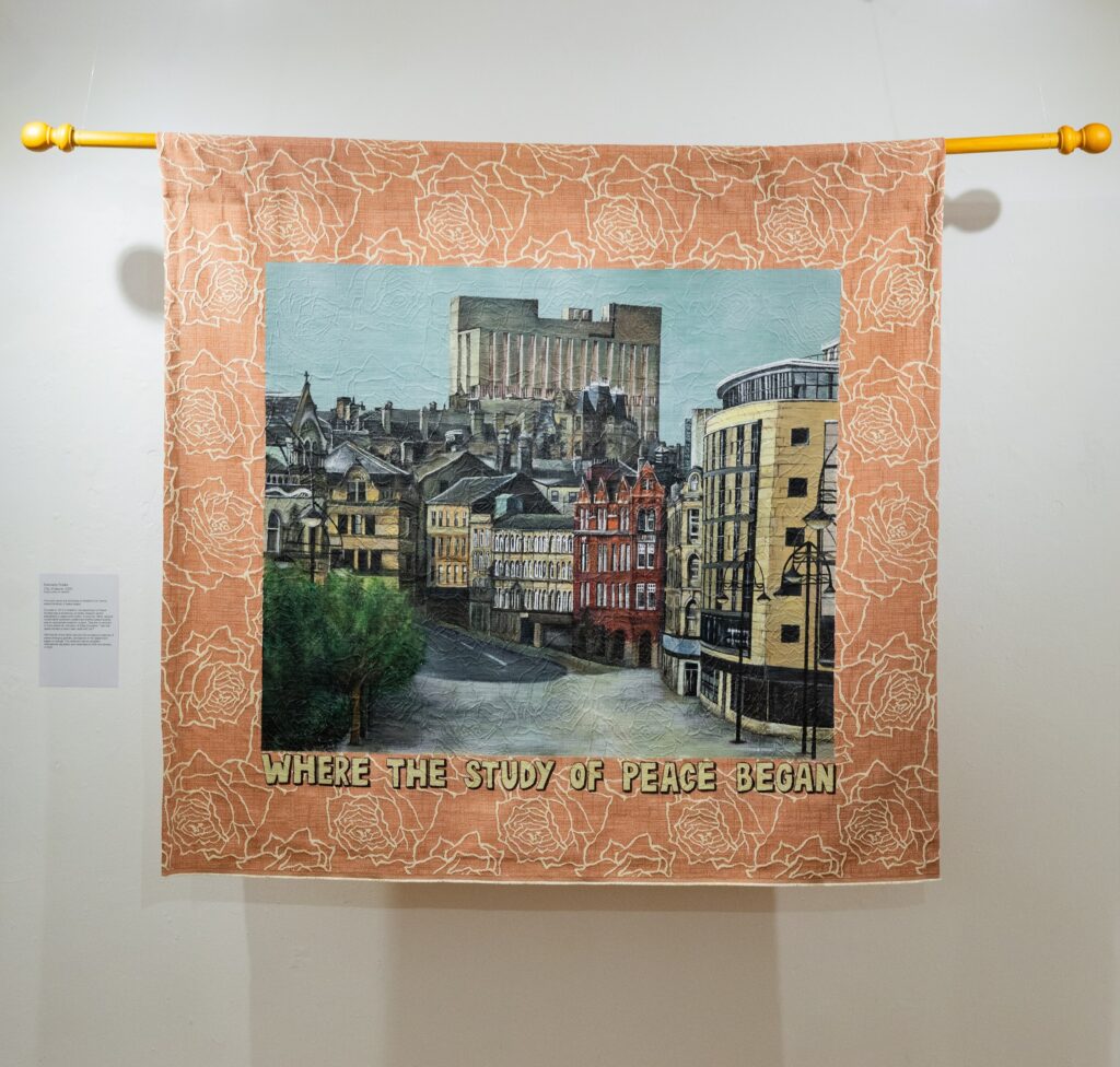 A hanging textile artwork showing a detailed stitched cityscape bordered by a patterned fabric frame, with the words “WHERE THE STUDY OF PEACE BEGAN” embroidered at the bottom.
