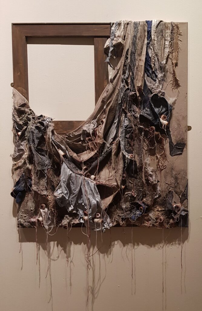 A mixed‑media wall artwork showing an empty wooden frame draped with torn, layered fabric pieces in earthy, worn textures.
