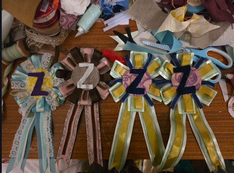 Four handmade ribbon rosettes on a craft table, with various colourful ribbons and fabric scraps scattered around.