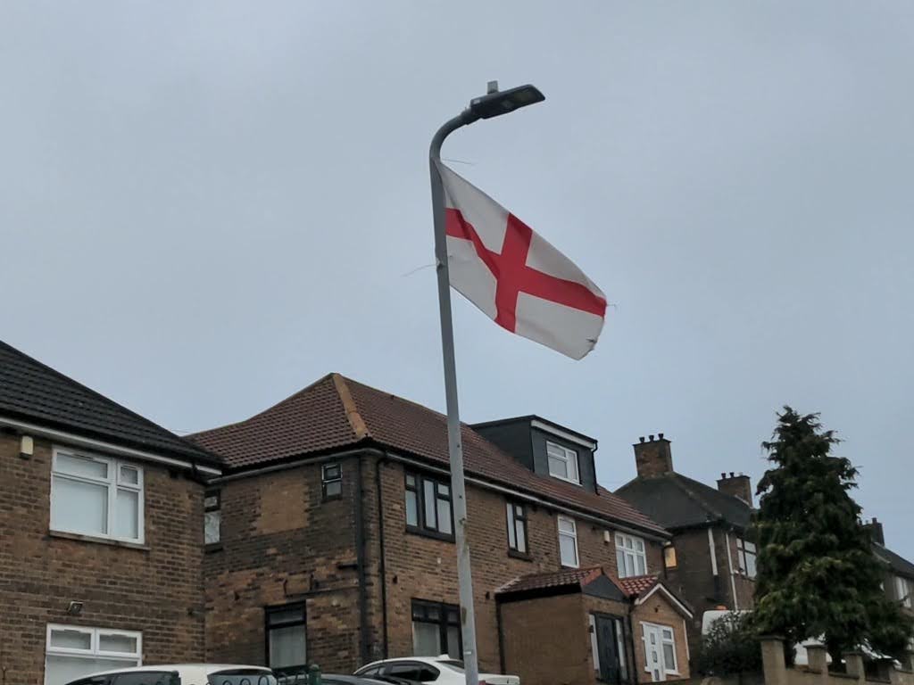 A St George’s flag mounted on a tall lamppost, blowing in the wind above a row of brick houses on a cloudy day.
