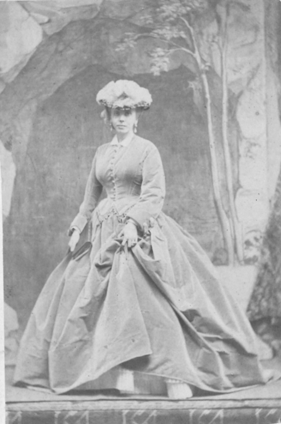 A 19th‑century studio portrait of a person wearing a voluminous Victorian dress with a fitted bodice and full skirt. They stand in front of a painted backdrop of trees and rocks, holding the sides of the dress. A large hat decorated with flowers completes the formal outfit.