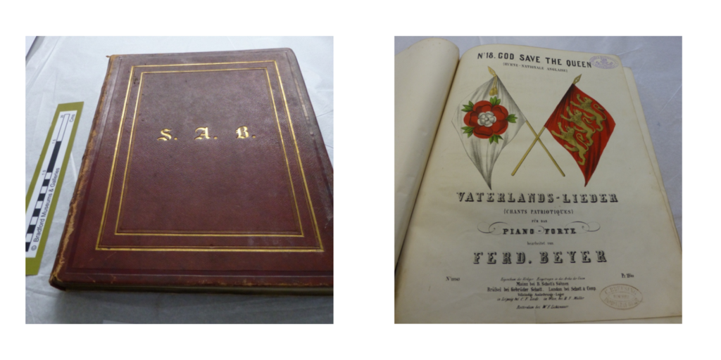 Here’s a clear, concise, and context‑appropriate alt‑text option for the image you uploaded:
Alt text:
Two 19th‑century music books belonging to Sarah Anna. One is a closed brown volume with gold trim and the initials “S.A.B.” on the cover. The other is open to a piece titled “Vaterlands‑Lieder… No. 18. God Save the Queen,” decorated with flags and a floral emblem.