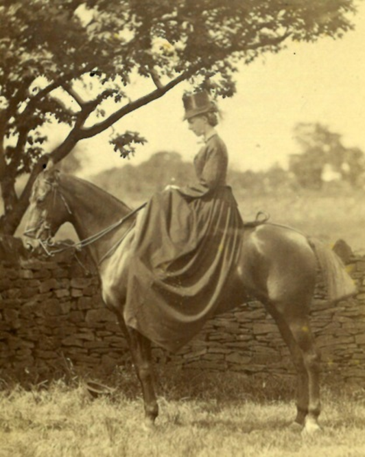 A sepia photograph of a person sitting side‑saddle on a horse in a grassy area. They wear a long Victorian dress and a hat, posed calmly beside a stone wall with a tree casting partial shade. Fields and trees stretch into the background.