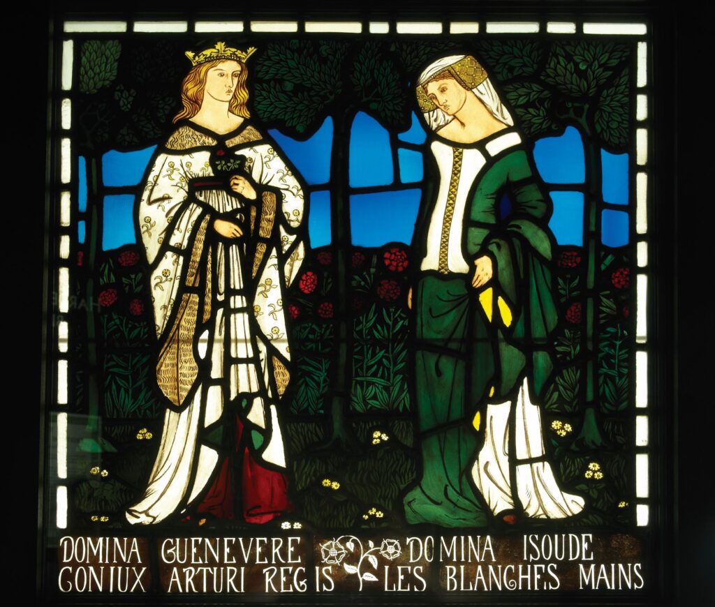 Stained‑glass panel depicting two women in a garden. On the left, a crowned woman in a white robe with gold details holds a red flower. On the right, a woman in a green dress with a white head covering stands among stylized trees and red flowers. Text below identifies them as Lady Guinevere, wife of King Arthur, and Lady Isoude of the White Hands.


