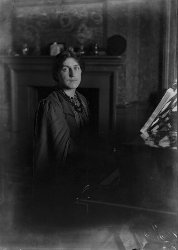  monochrome portrait showing a person seated indoors near an ornate fireplace. The individual is dressed in dark, formal clothing with layered fabric and a beaded necklace. The room has patterned wallpaper and decorative objects arranged on the mantelpiece. To the right, a piano or similar instrument is partially visible with a sheet‑music stand holding open pages. The overall photograph has a soft, shadowed quality.