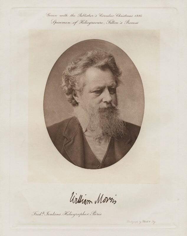 A sepia‑toned oval portrait mounted on a cream paper background. The photograph shows the upper body of an older individual with curly hair and a long, textured beard, wearing a collared shirt, waistcoat, and jacket. Above the portrait, printed text reads: “Given with the Publisher’s Circular Christmas 1885 / Specimen of Heliogravure, Gillot’s Process.” Below the portrait is a handwritten signature that reads “William Morris,” with small printed text beneath indicating the photographer’s studio in Paris.