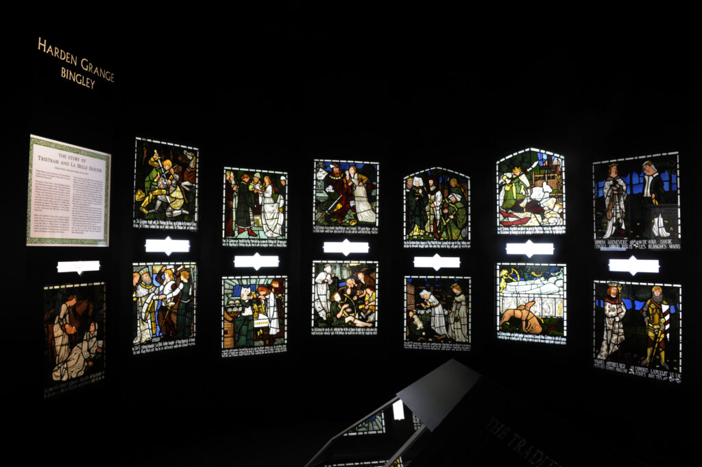 A display of illuminated stained‑glass panels arranged in two rows, each depicting scenes from the medieval romance of Tristram and La Belle Isolde. To the left is a framed text panel summarizing the legend. Above the glass panels, the words “HARDEN GRANGE BINGLEY” appear. The colours are vivid against a dark background, highlighting the intricate designs.