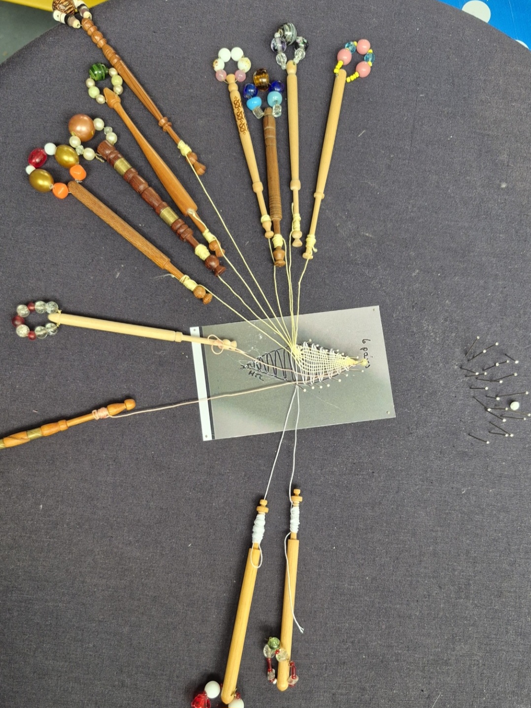 A bobbin‑lace pillow with multiple wooden bobbins arranged around a small rectangular pattern card. Fine threads are crossed and pinned into the printed design, with extra pins gathered to the side. The bobbins have decorative beads, and the setup rests on dark fabric.