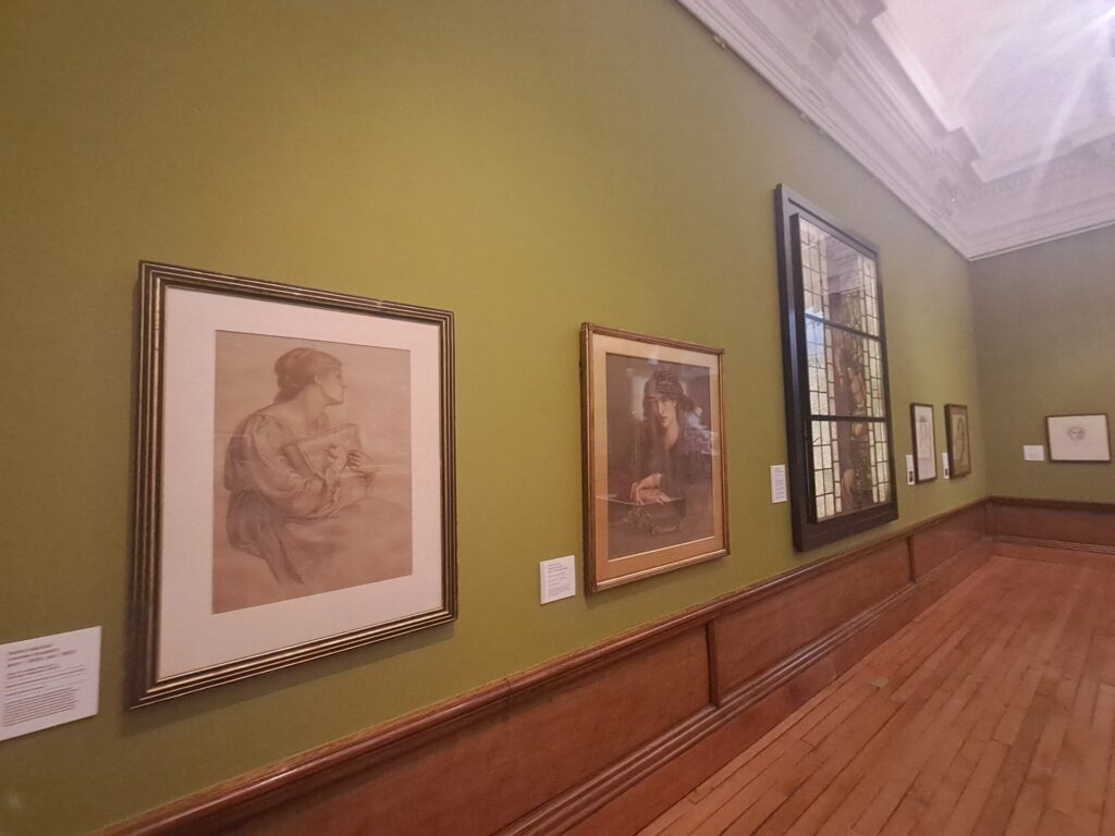A gallery interior with a row of framed artworks displayed on a green wall. The works include portraits and sketches and stained glass each with a small wall label. The space has wooden floors and an ornate white ceiling, giving a traditional museum setting.