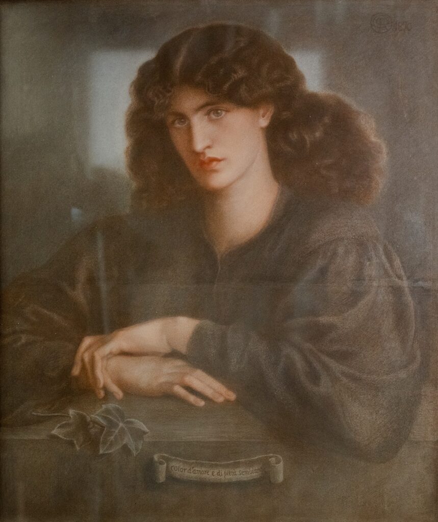 A painted half‑length portrait of a person with long wavy brown hair, seated with hands loosely crossed on a surface. A small plant and a scroll with Italian text sit in the foreground. The background is softly blurred, drawing attention to the calm, direct gaze of the sitter.