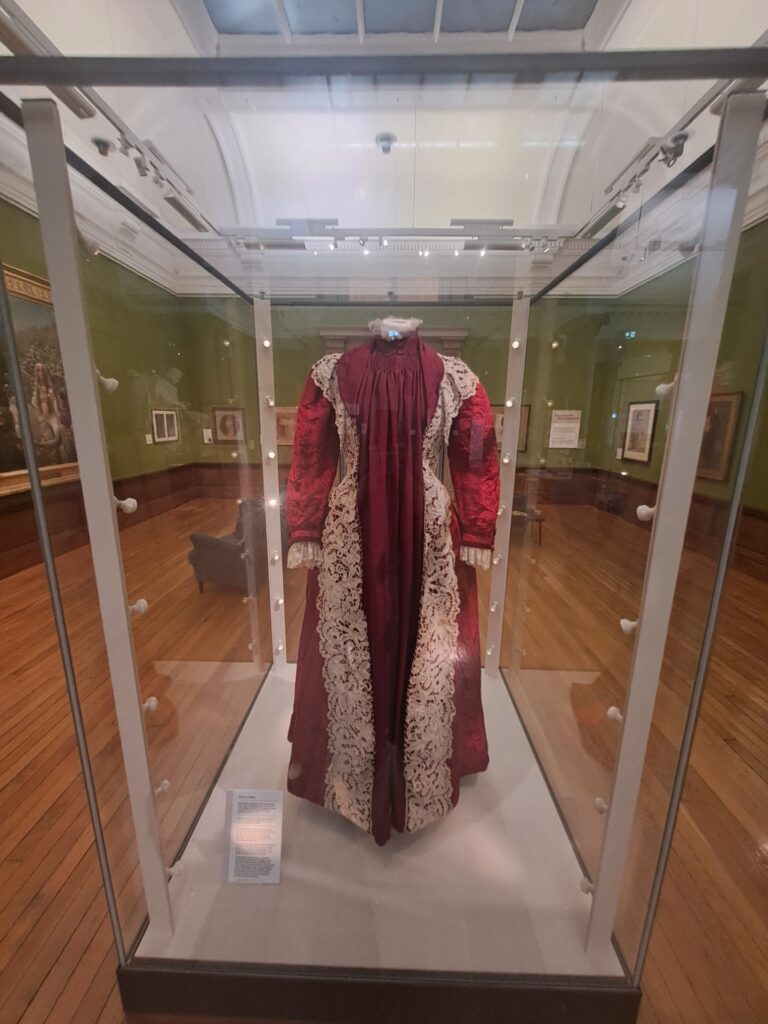 burgundy dress displayed on a mannequin inside a glass museum case. The floor-length gown features elaborate cream lace trim running down the front, around the cuffs, and along the high collar. The lighting highlights the rich texture of the fabric and lace