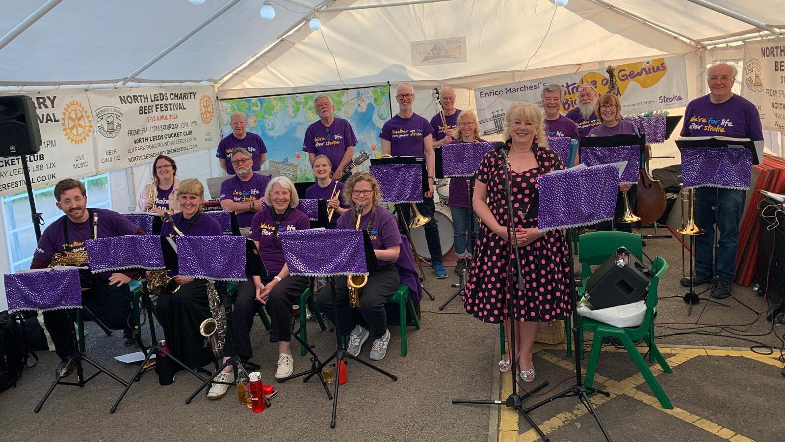 Big band in purple Stroke Association shirts performing under a white tent with brass instruments and a vocalist in front.